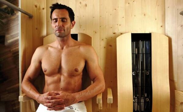 Un hombre de pie en una habitación de madera, con la puerta de una sauna y un banco de madera.