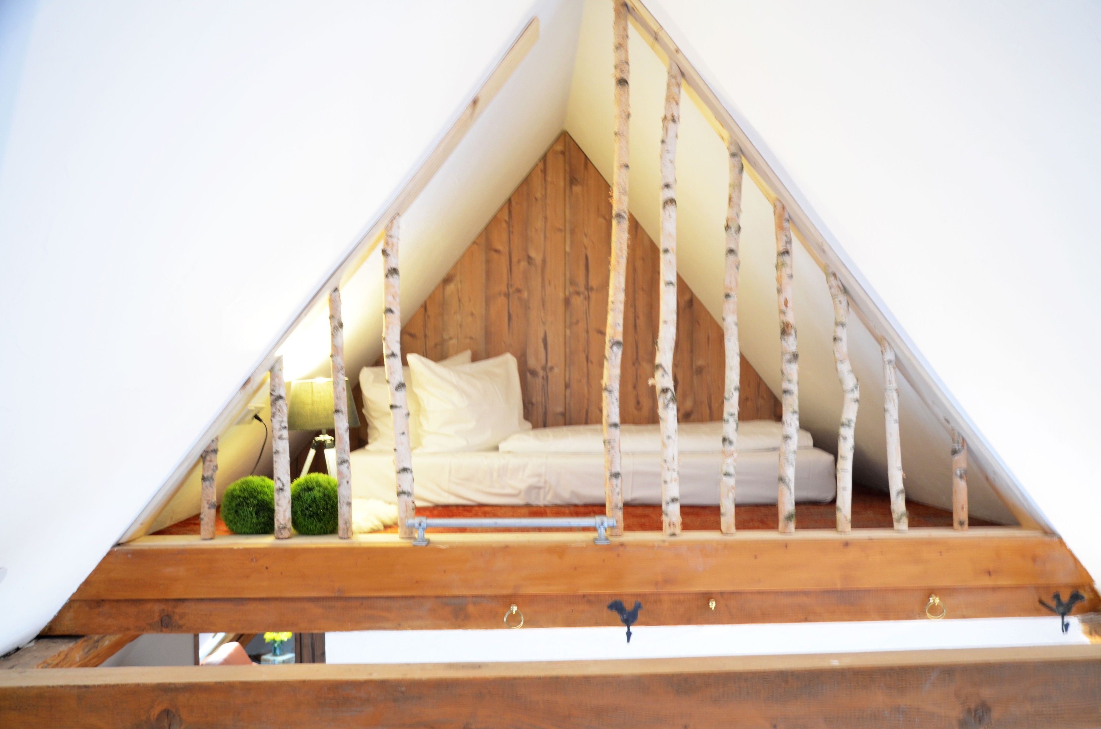 A triangular attic bedroom with a wooden floor, a bed with white bedding, and birch tree branches as a decorative railing.