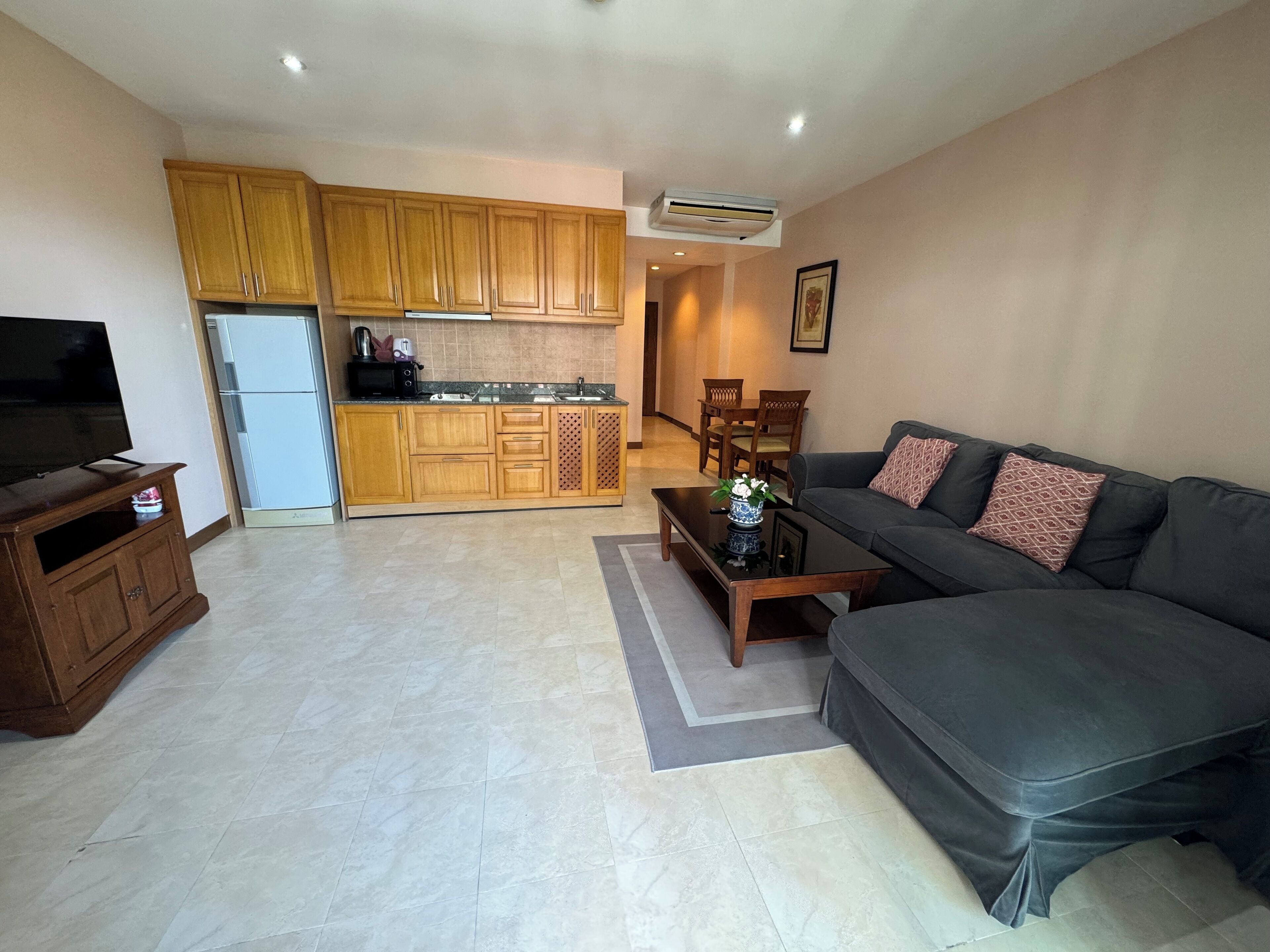 A living room with a sofa, a coffee table, and a TV on a wooden cabinet. There is a kitchen area with wooden cabinets and a refrigerator.