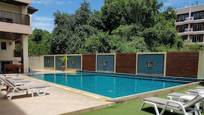 A swimming pool with lounge chairs, surrounded by a green lawn and a building with a tiled wall and wooden panels. - Jomtien Beach Residence (Pattaya)