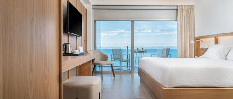 A hotel room with a large bed, a desk with a flat-screen TV, a chair, and a view of the sea.