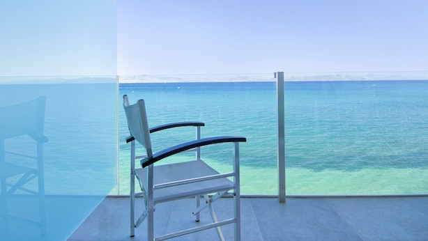 A chair facing a clear blue sea from a balcony with a glass barrier. - Kos Aktis Art Hotel (Kos)