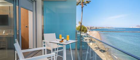 A balcony with white chairs and a table, a palm tree, and a view of the sea and distant buildings.
