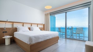 A modern hotel room with a large bed, a bedside table with a lamp, a small table with chairs, and a balcony with a view of the sea. - Kos Aktis Art Hotel (Kos)