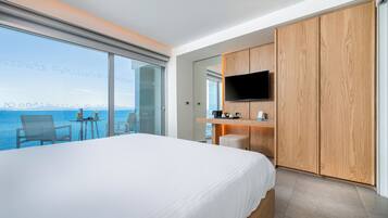 A hotel room with a large bed, a TV mounted on a wooden cabinet, a small table with a chair, and a view of the sea.