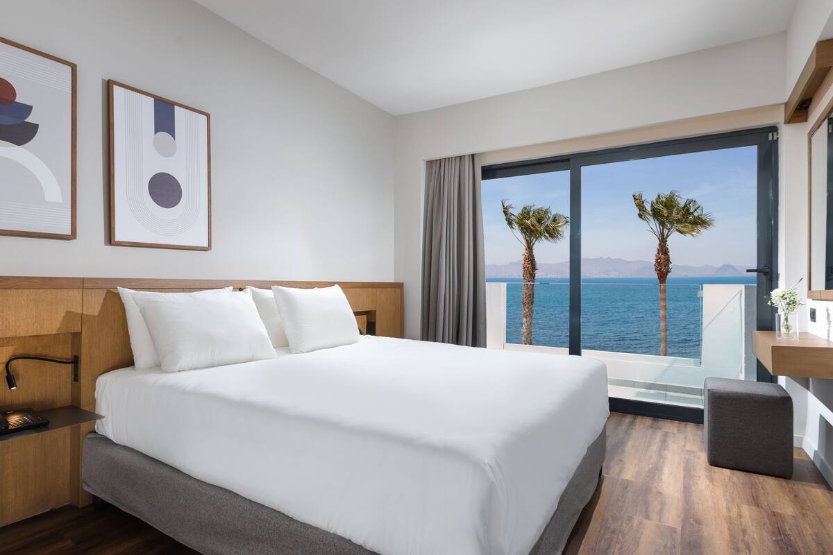 A modern hotel room with a large bed, a wooden headboard, a desk with a lamp, and a view of the sea with palm trees.