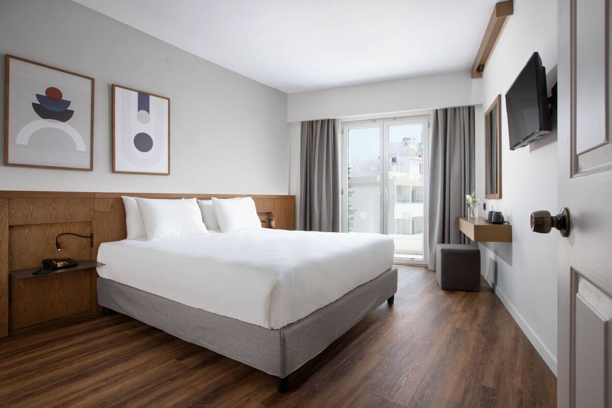A hotel room with a large bed, wooden headboard, a bedside table with a lamp, a TV mounted on the wall, and a sliding glass door leading to a balcony.