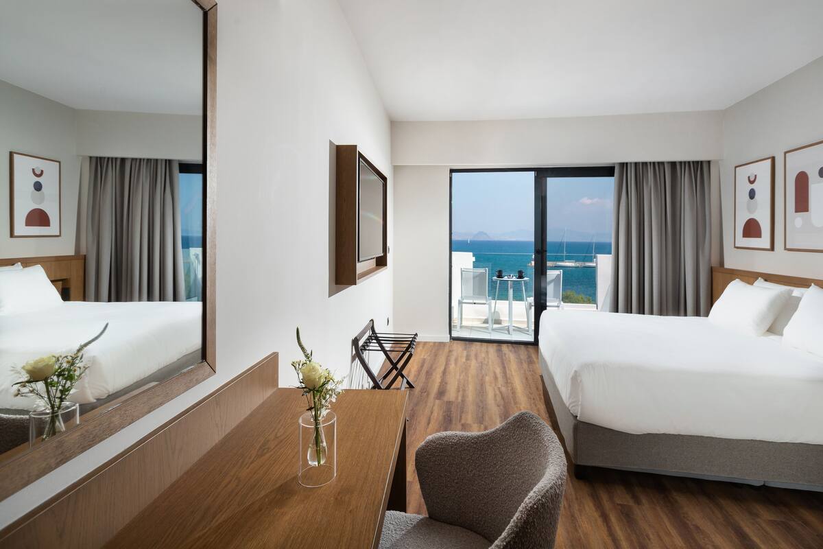A hotel room with two beds, a wooden dining table with a vase of flowers, and a large window offering a view of the sea and boats.