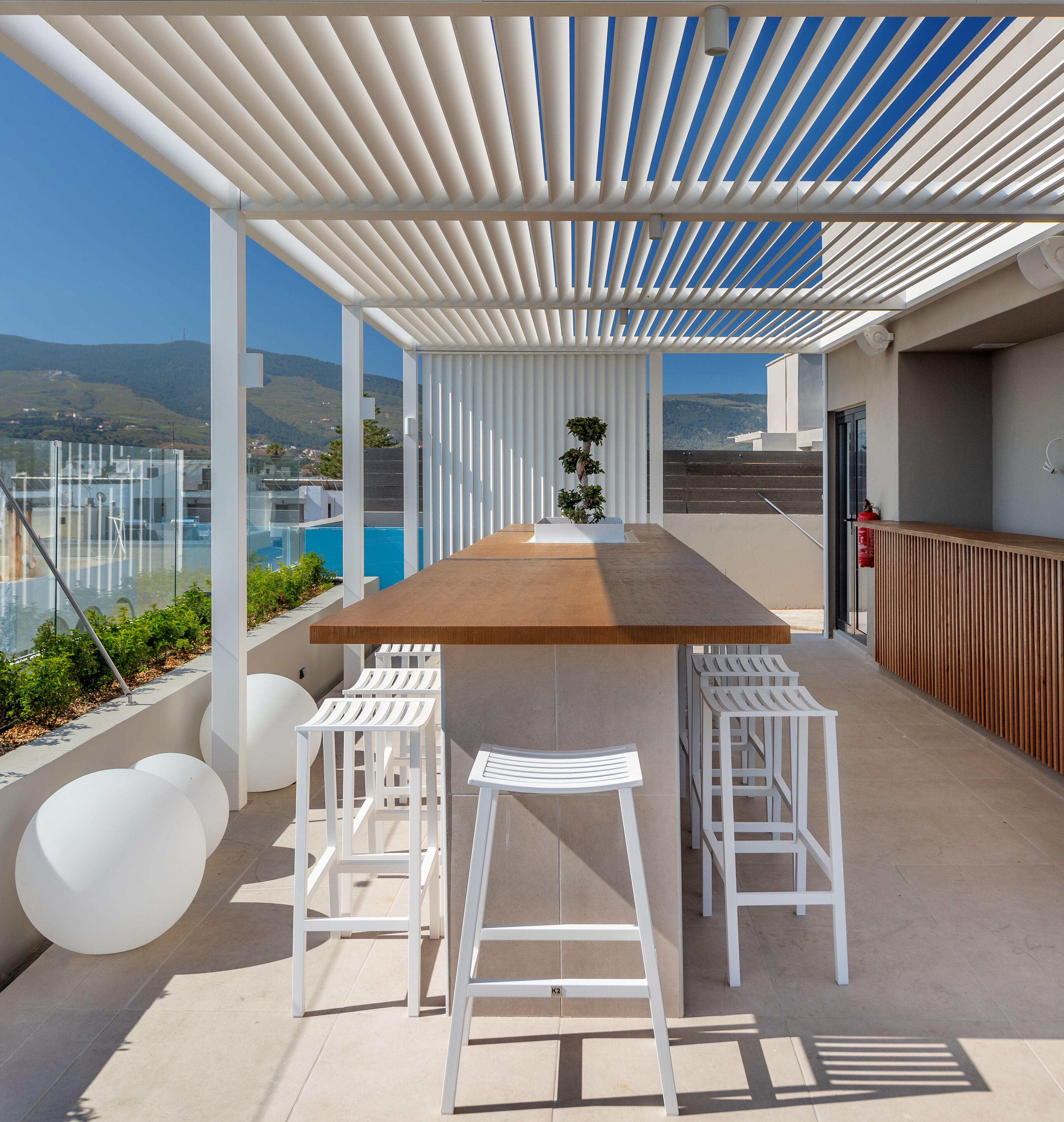 A modern outdoor patio with a wooden table and white bar stools, overlooking a pool and mountains.