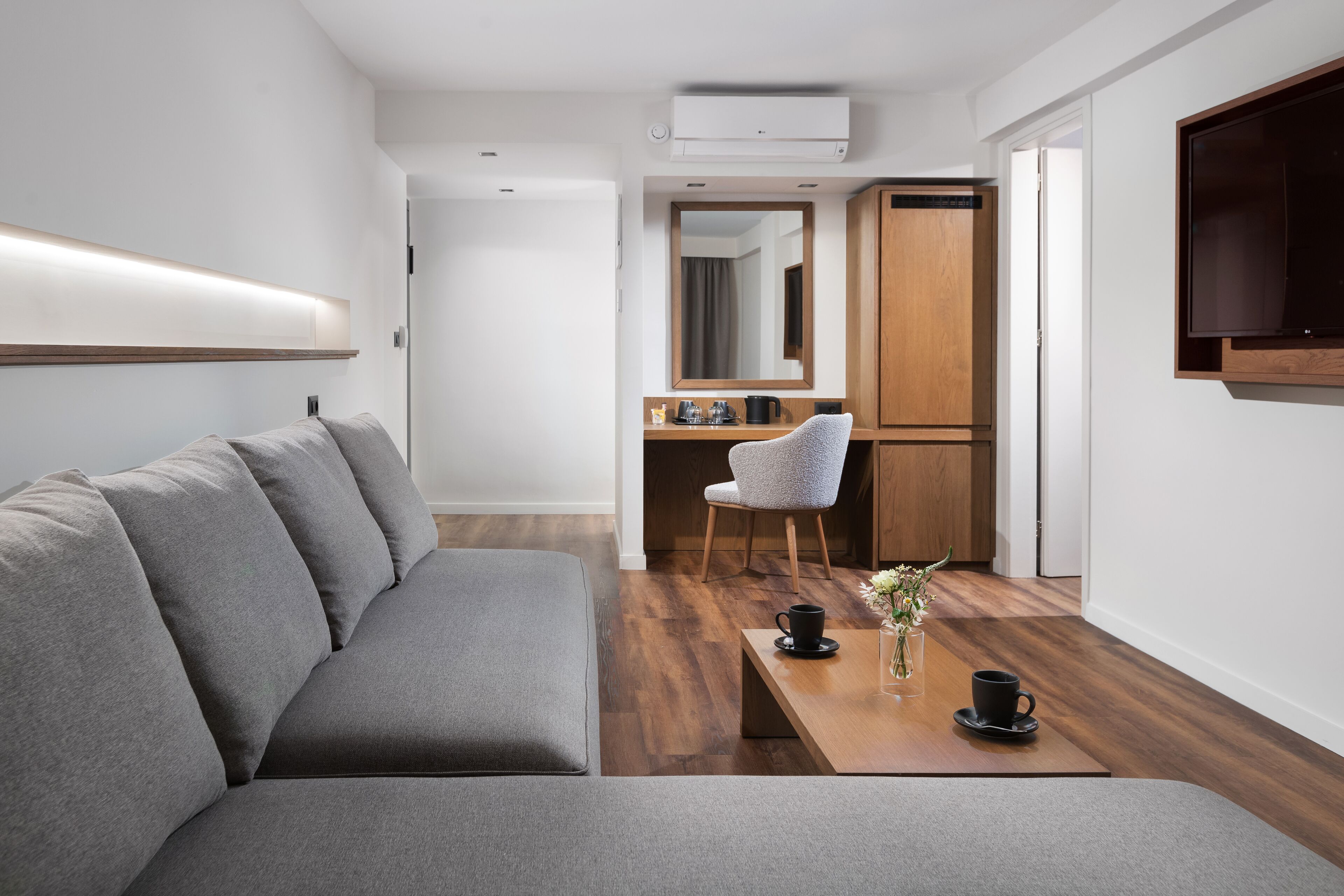 A modern living room with a grey sofa, a wooden coffee table, and a small dining area with a chair and a mirror.