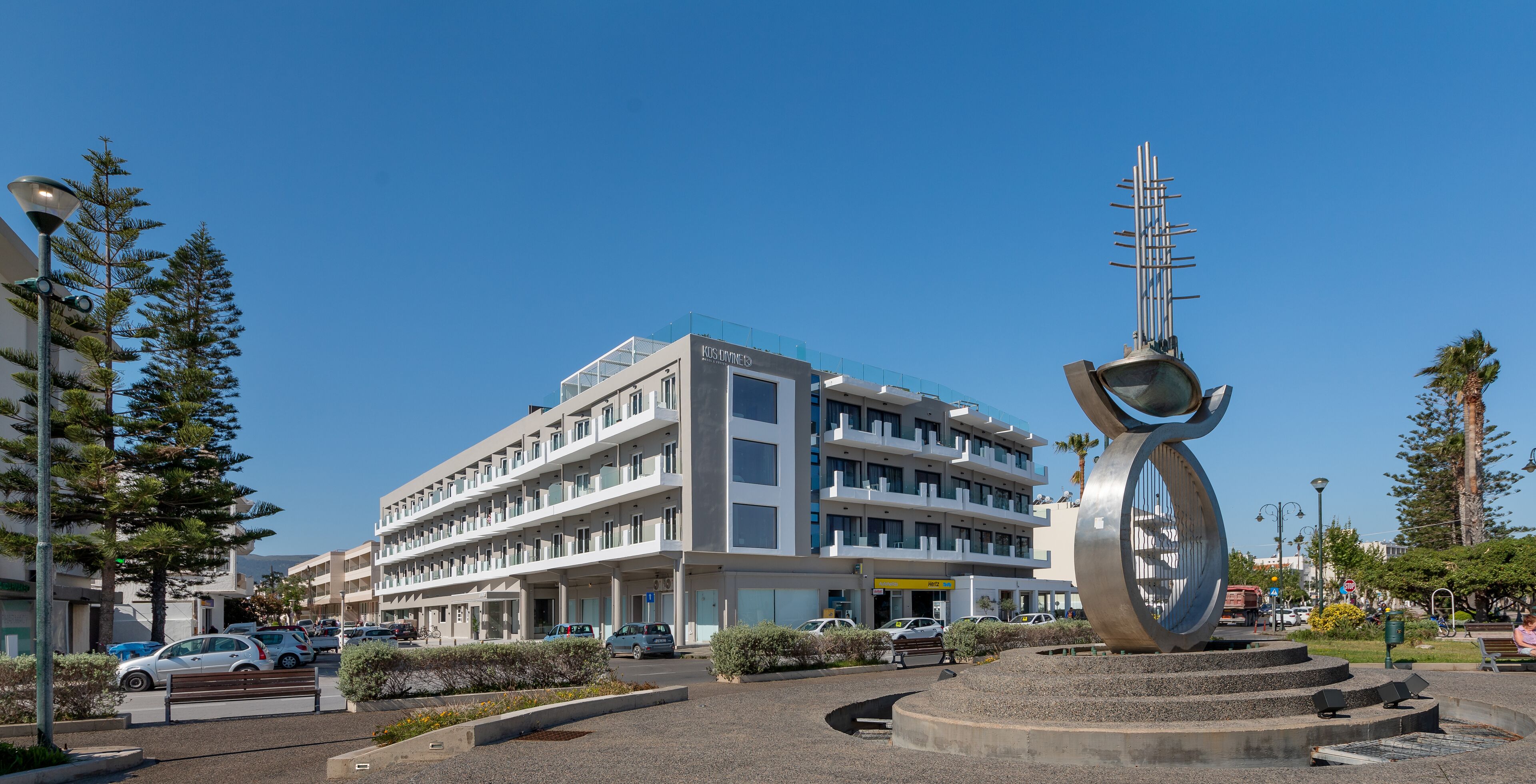 Un edificio moderno de varios pisos con un atrio central, rodeado de un área ajardinada con bancos y una gran escultura de metal.