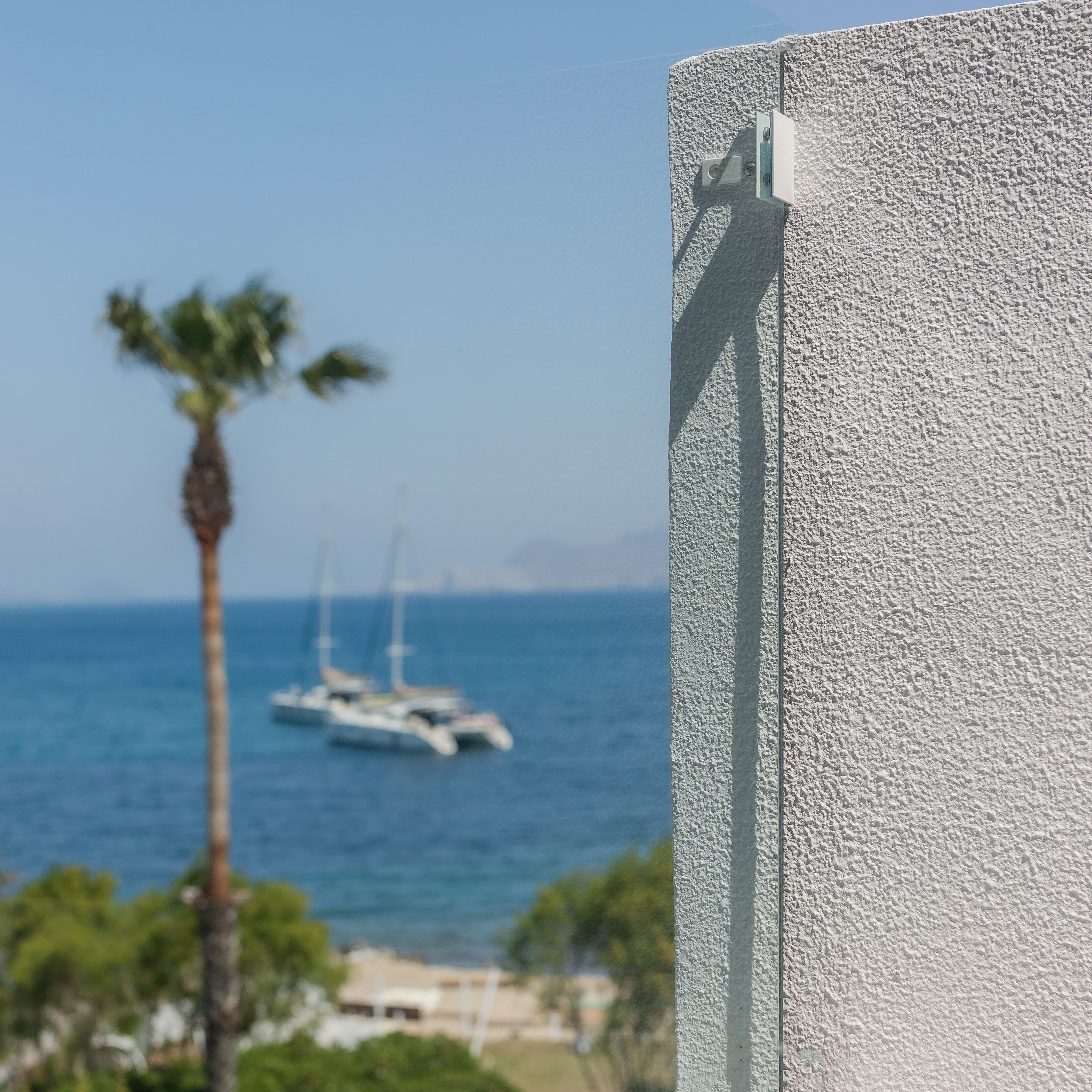 Un mur à la texture particulière, agrémenté d’un support métallique, d’un palmier et de voiliers sur la mer.