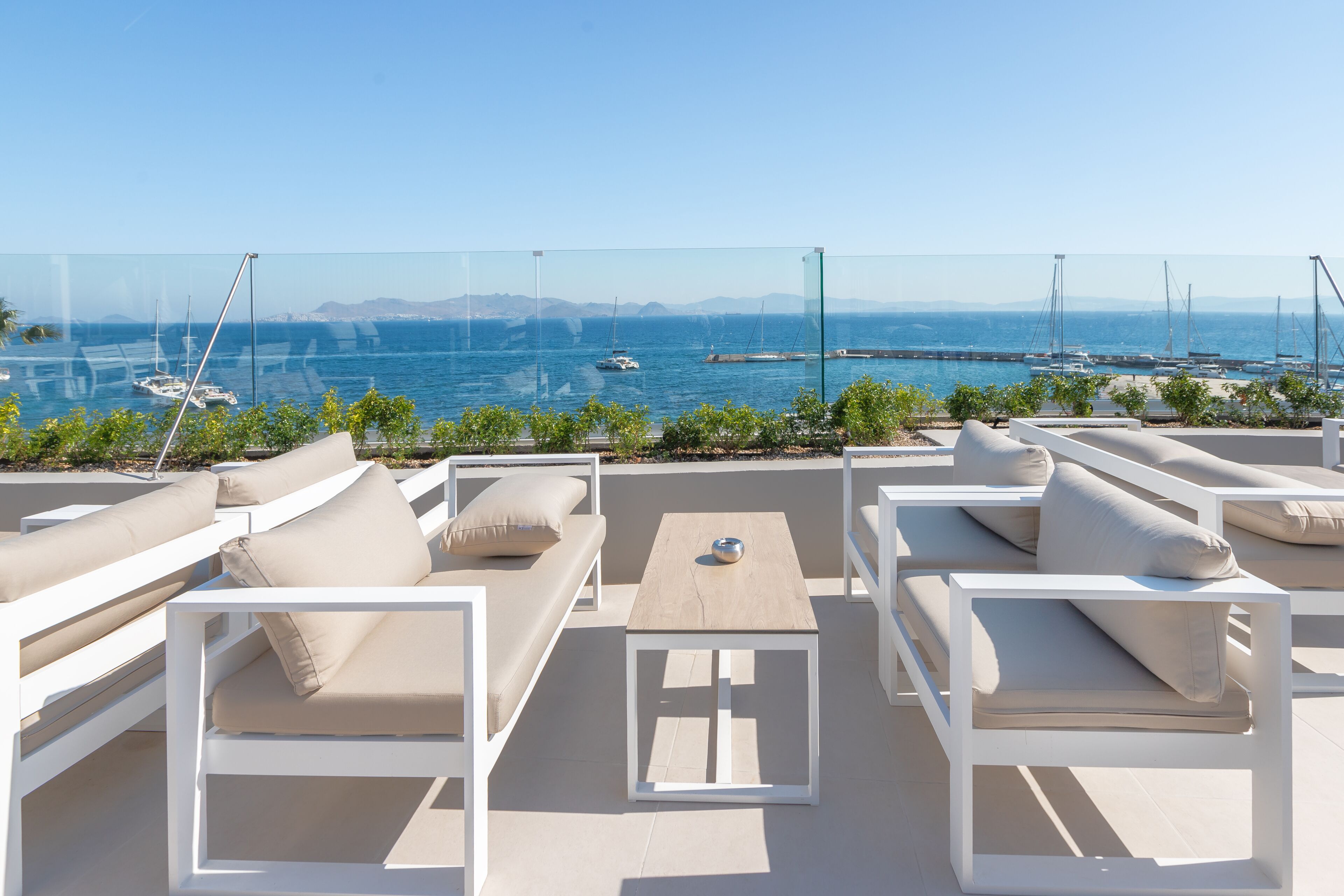 Terraza en la azotea con mobiliario blanco, una mesa de madera y vistas al mar y barcos.