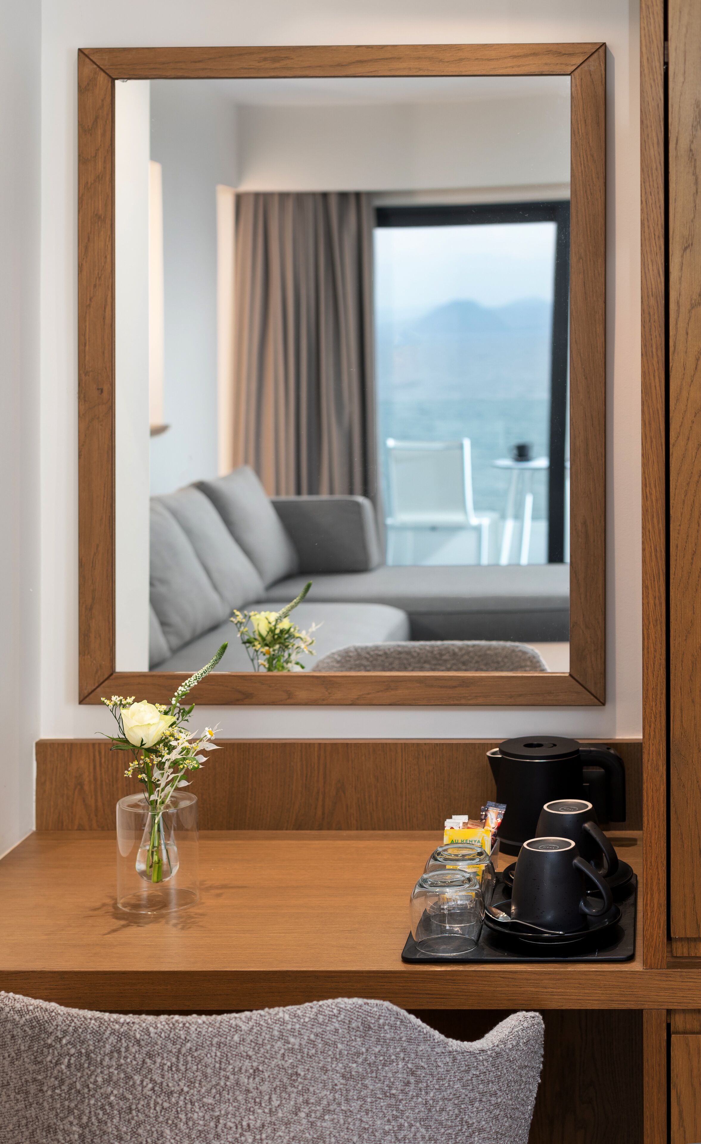 Une chambre d’hôtel avec un bureau en bois, un vase de fleurs, une machine à café et une vue sur un balcon agrémenté d’un canapé et de rideaux.