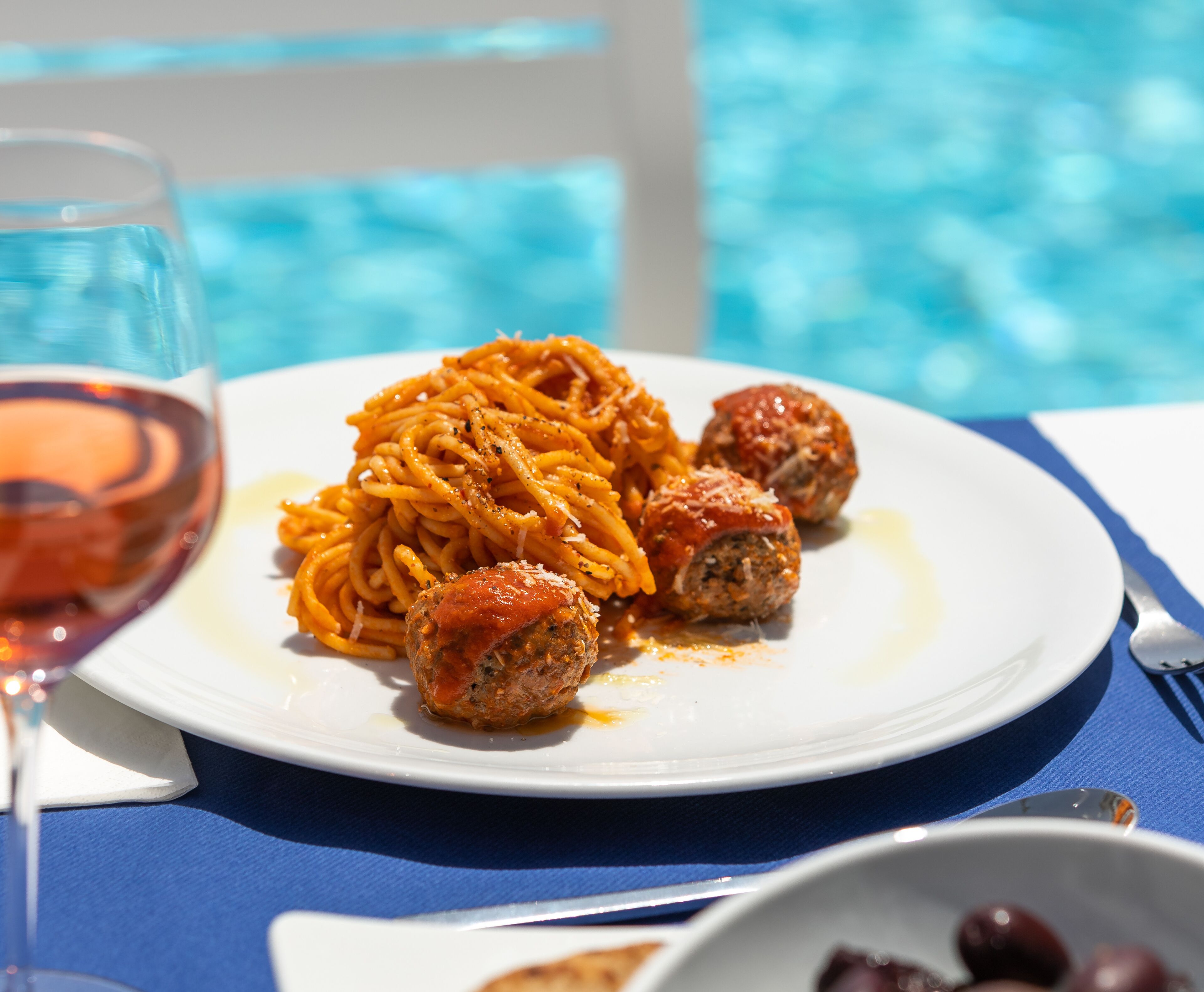 A plate of spaghetti and meatballs with a glass of red wine on a table by a pool.
