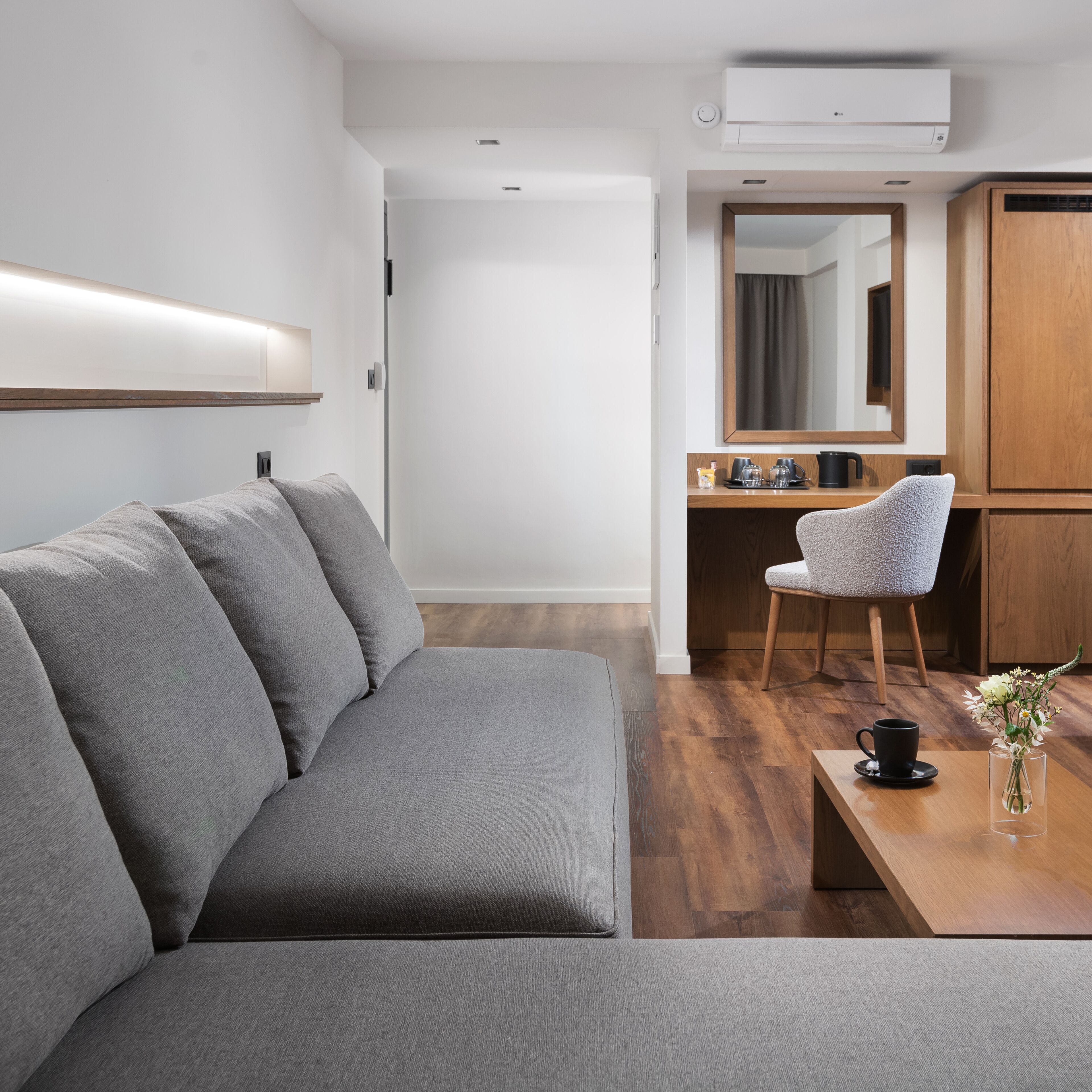 A modern hotel room with a grey sofa, a wooden desk, a chair, a coffee table, and a vase with flowers.