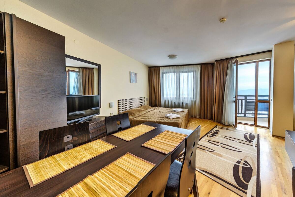 Una habitación de hotel con cama, televisión, mesa de comedor con sillas, balcón con cortinas y vistas al exterior.