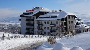 A multi-story hotel with a snow-covered roof and balconies, surrounded by a snowy landscape and mountains in the background. - All Seasons Club (Bansko)