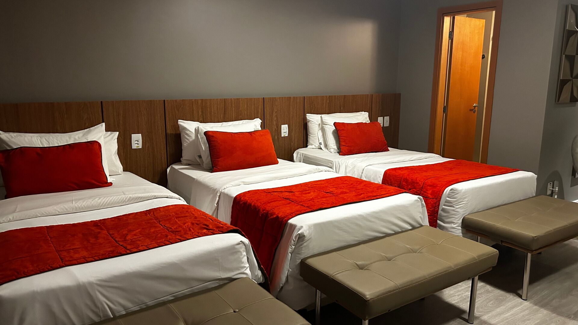 Habitación de hotel con cuatro camas, cada una con ropa de cama blanca y detalles rojos. Hay dos mesitas de noche y un banco en el centro.