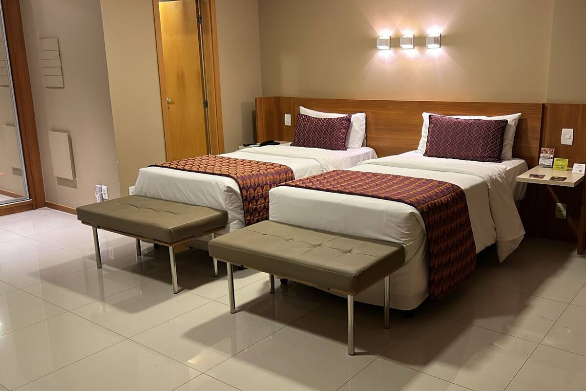 A hotel room with two beds, a small table, and a bench.