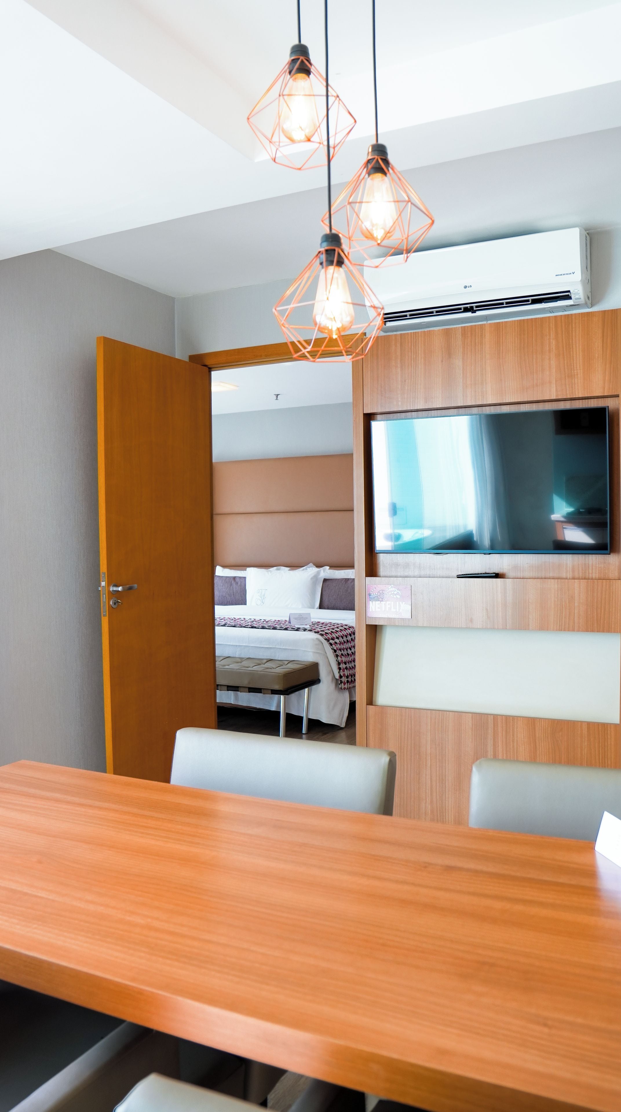 Una habitación de hotel moderna con una mesa de comedor de madera, una cama con cabecero, un televisor montado en la pared y aire acondicionado en el techo.