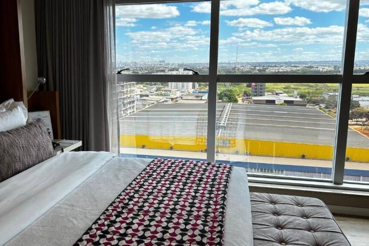A hotel room with a large window, a bed with a patterned bedspread, a grey upholstered headboard, and a view of a sports field and buildings.