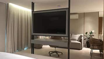 Quarto de hotel com cama, escrivaninha com cadeira, televisão em suporte, sofá e planta em vaso.