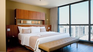 A hotel room with a large bed, a bench, a wooden headboard, a nightstand, and a view of buildings through a large window. - Jade Hotel Brasília (Guará)