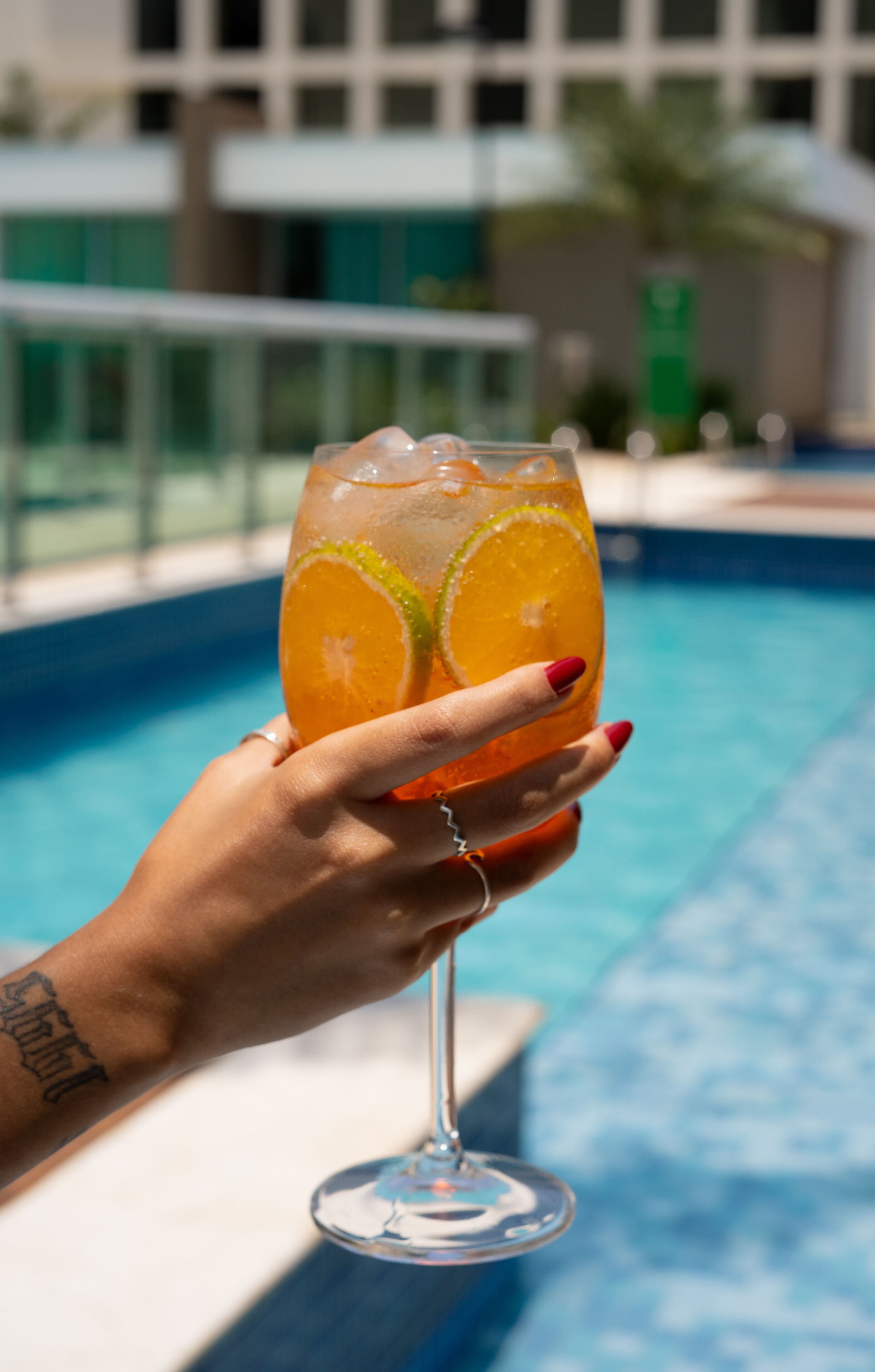 A hand holding a cocktail glass with a slice of orange and lime, near a swimming pool.