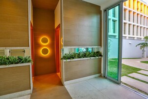 Un moderno vestíbulo de hotel con un mostrador de recepción de madera, una puerta de vidrio que da al exterior y una pequeña zona ajardinada con plantas.
