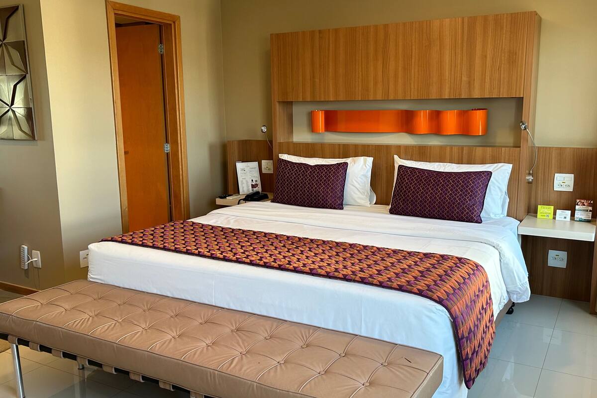 A hotel room with a large bed, a wooden headboard, a bedside table, a bench, and a bathroom with a purple chair.