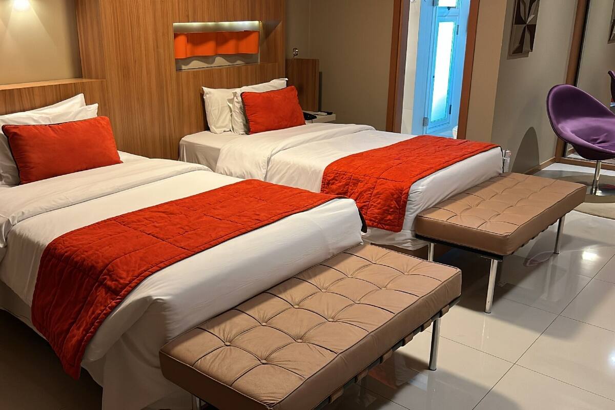 A hotel room with two beds, each with white bedding and orange accents, a bench, a purple chair, and a wooden headboard.