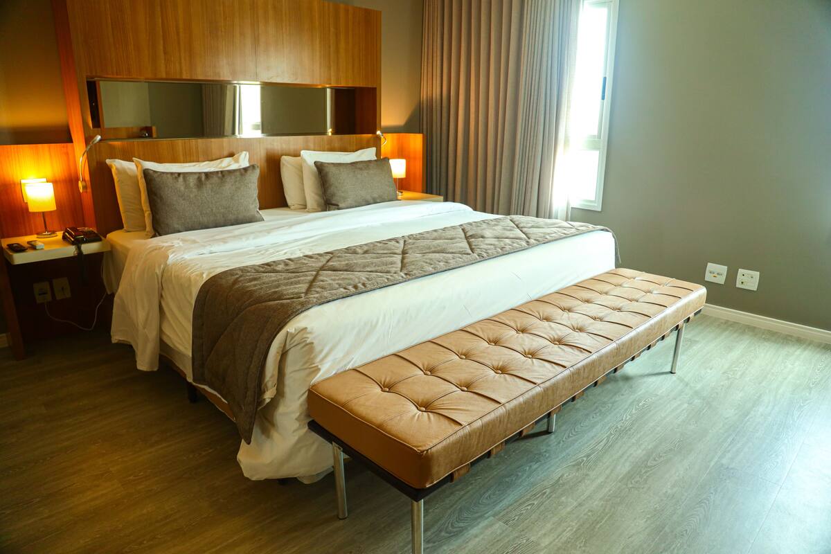 A hotel room with a large bed, a bedside table with a lamp, a bench at the foot of the bed, and a large mirror above the bed.
