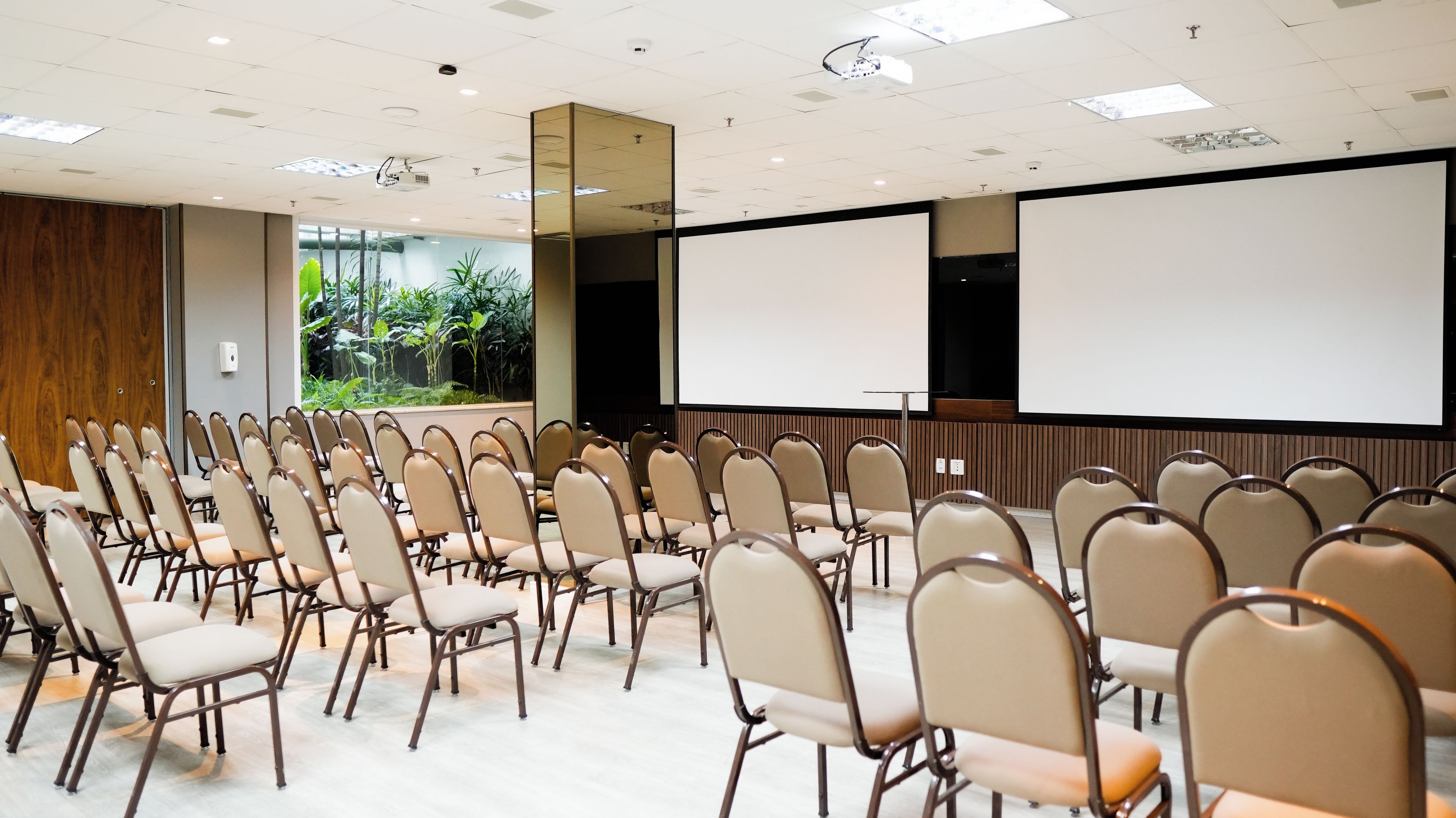 A conference room with rows of chairs, a projector screen, and a large window with a view of greenery.
