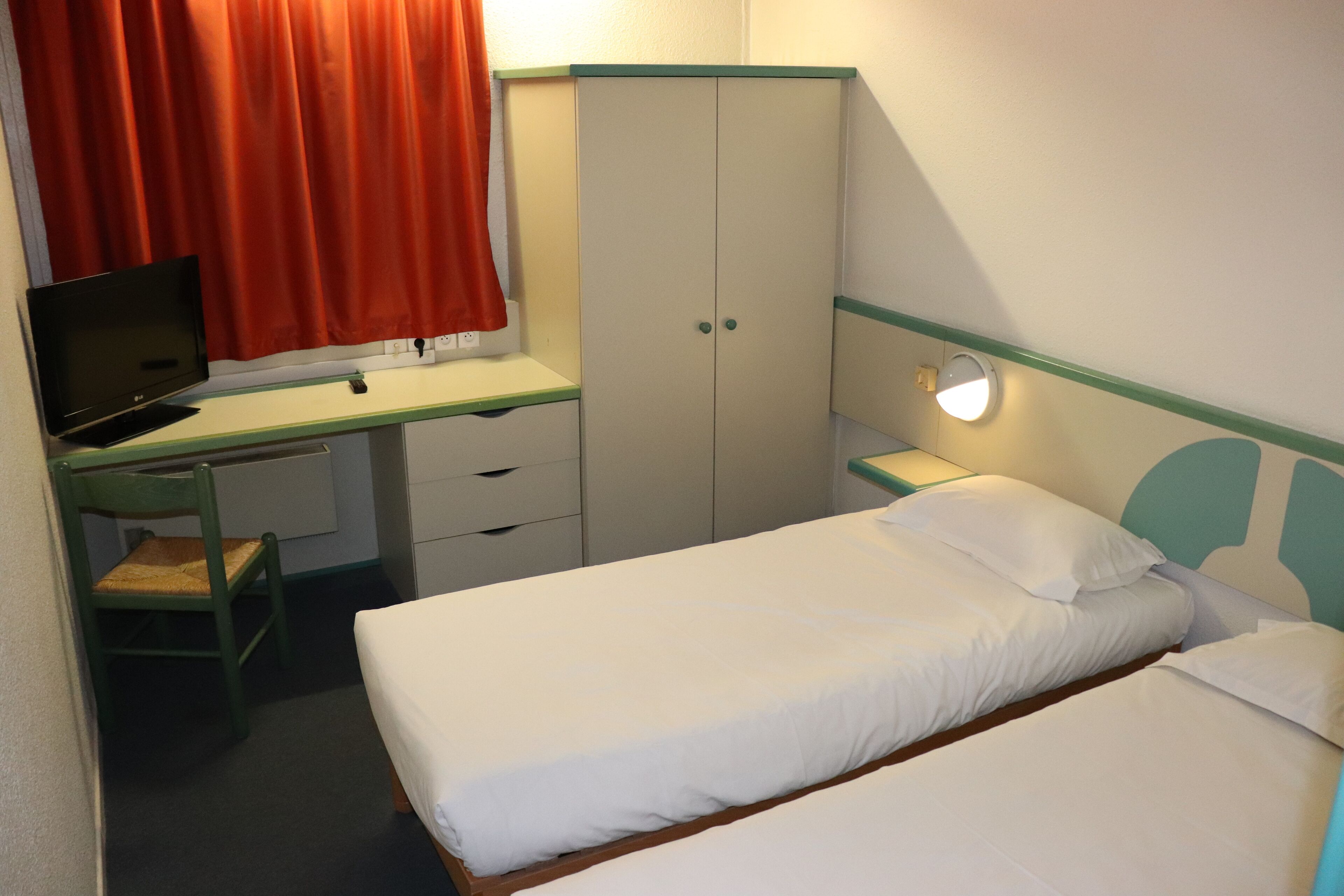 A small, neatly arranged bedroom with a single bed, a desk with a laptop, a wardrobe, and a bedside table with a lamp.