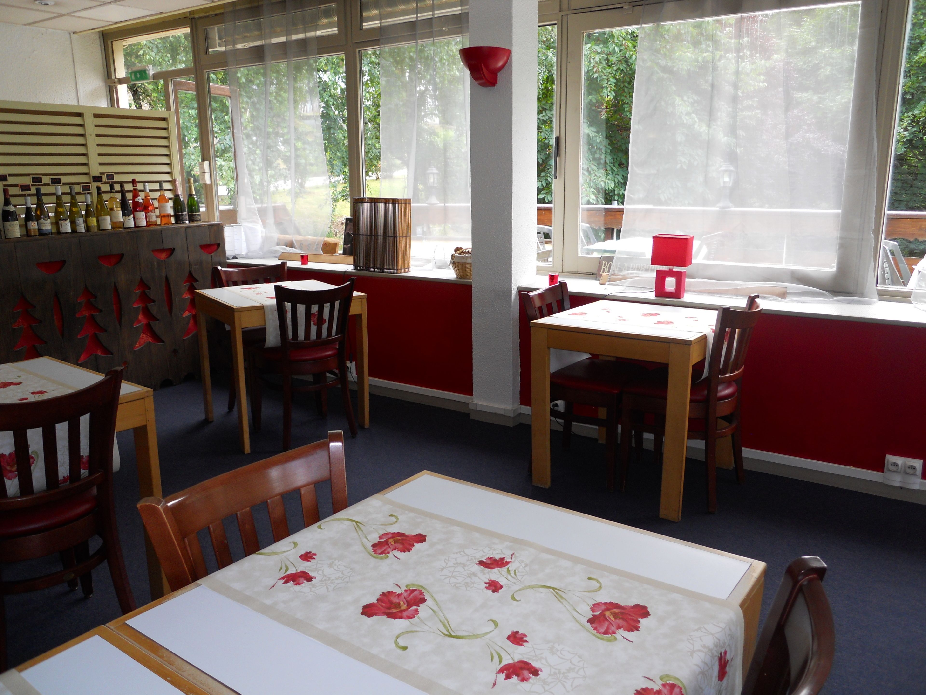 A dining area with wooden tables and chairs, a red wall, and a window with a view of greenery.