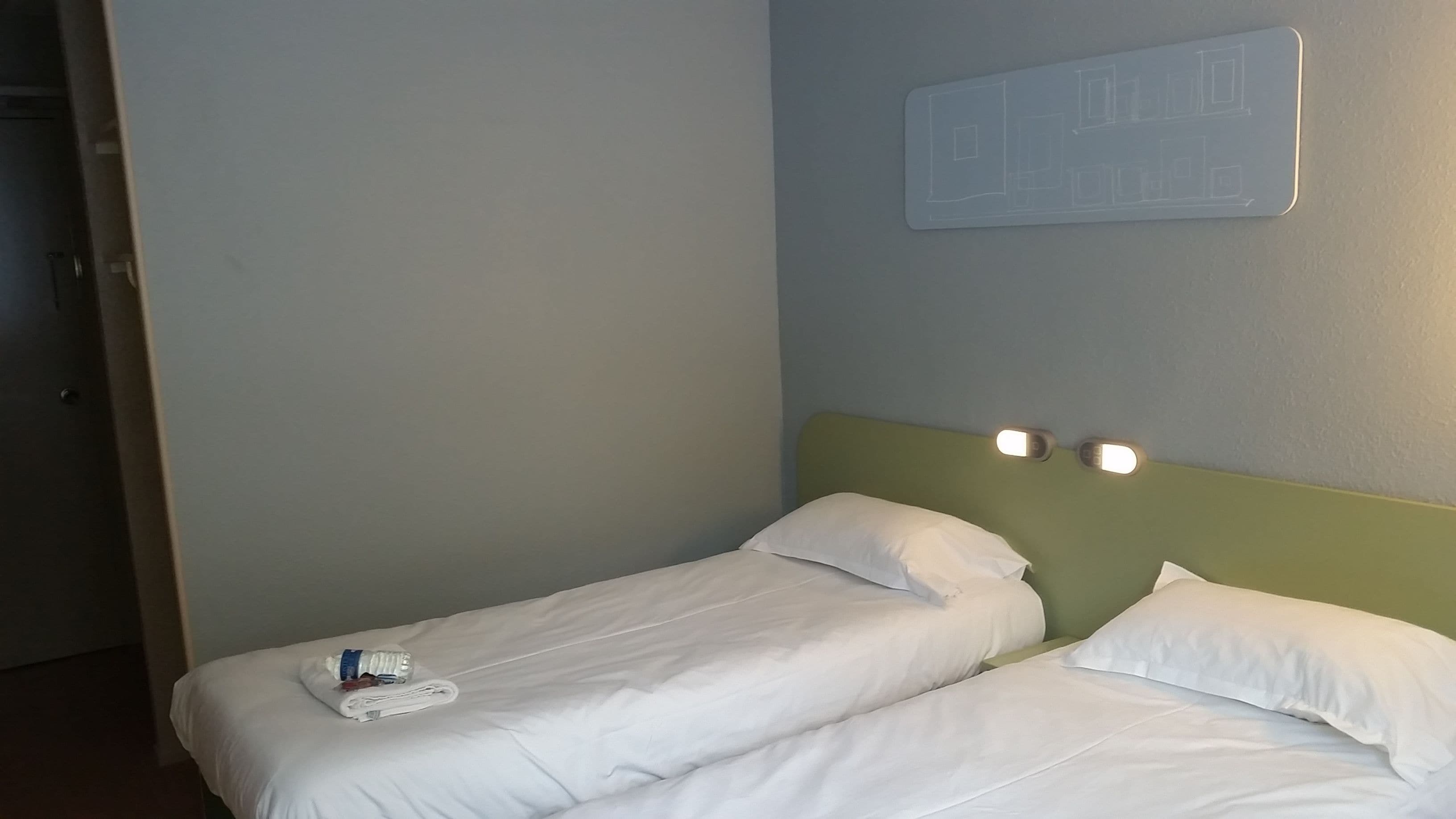 A hotel room with two single beds, a wall-mounted light switch, and a door.