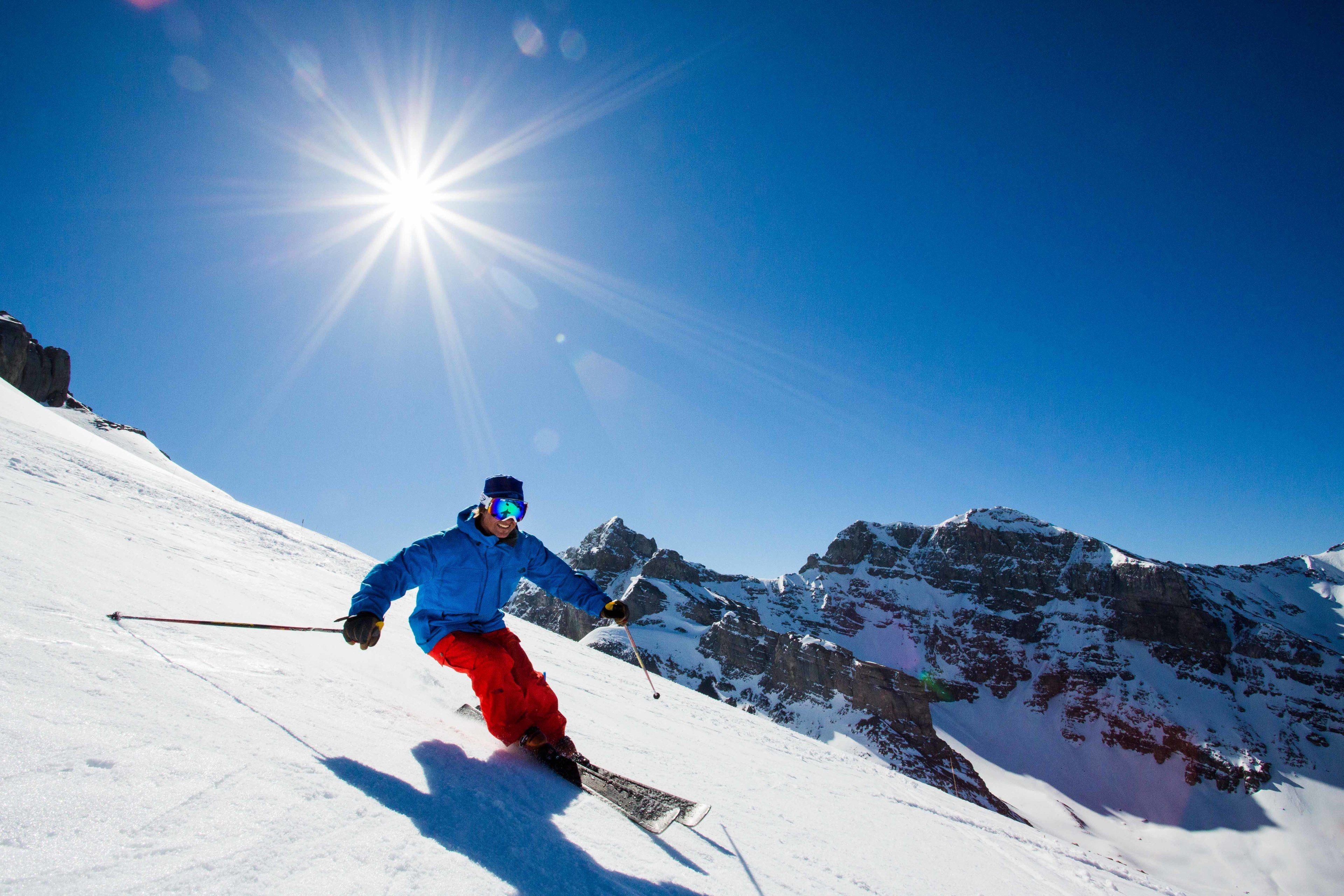 A person skiing down a snow-covered slope with a clear blue sky and sun in the background.