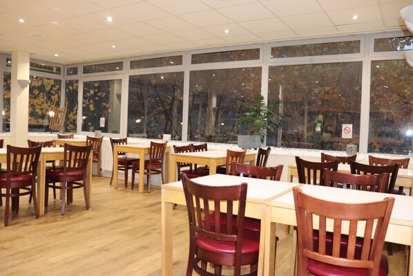 A restaurant with wooden tables and chairs, large windows with autumn leaves, and a potted plant.