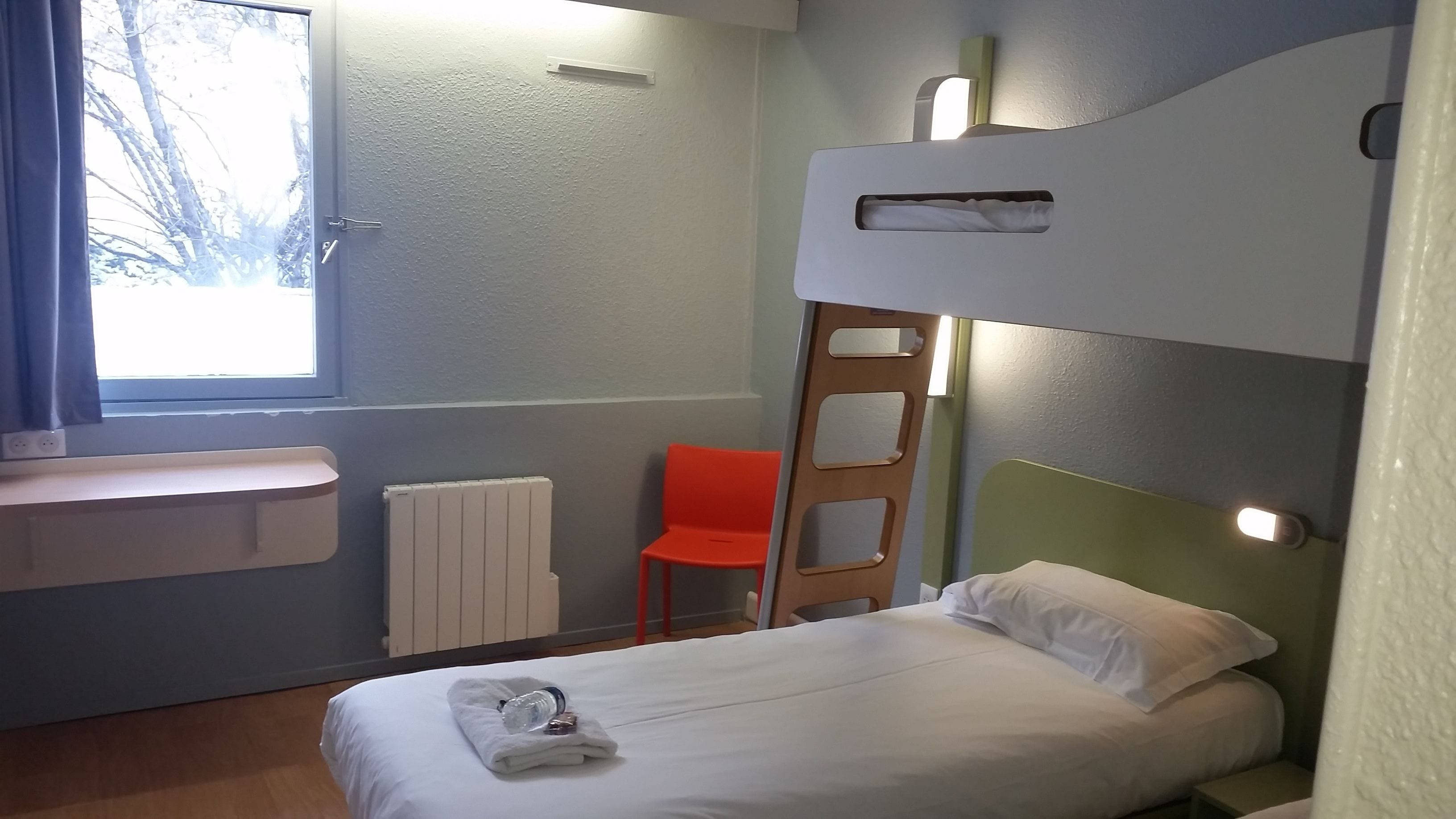 A small, neatly arranged room with a bunk bed, a single chair, a small table, and a window providing natural light.