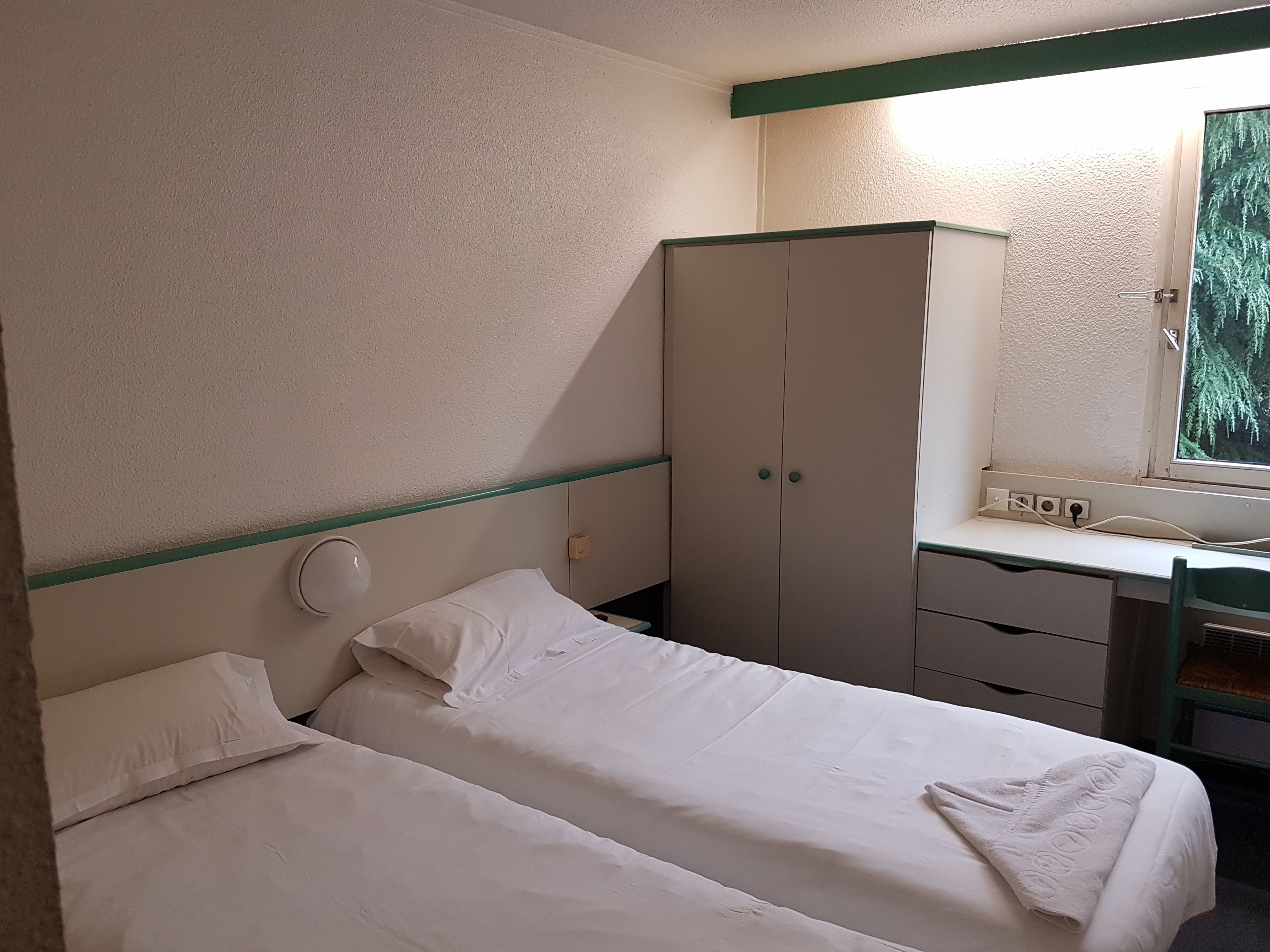 Ett hotellrum med två sängar, en garderob, ett skrivbord med en spegel och ett fönster med utsikt över träd.