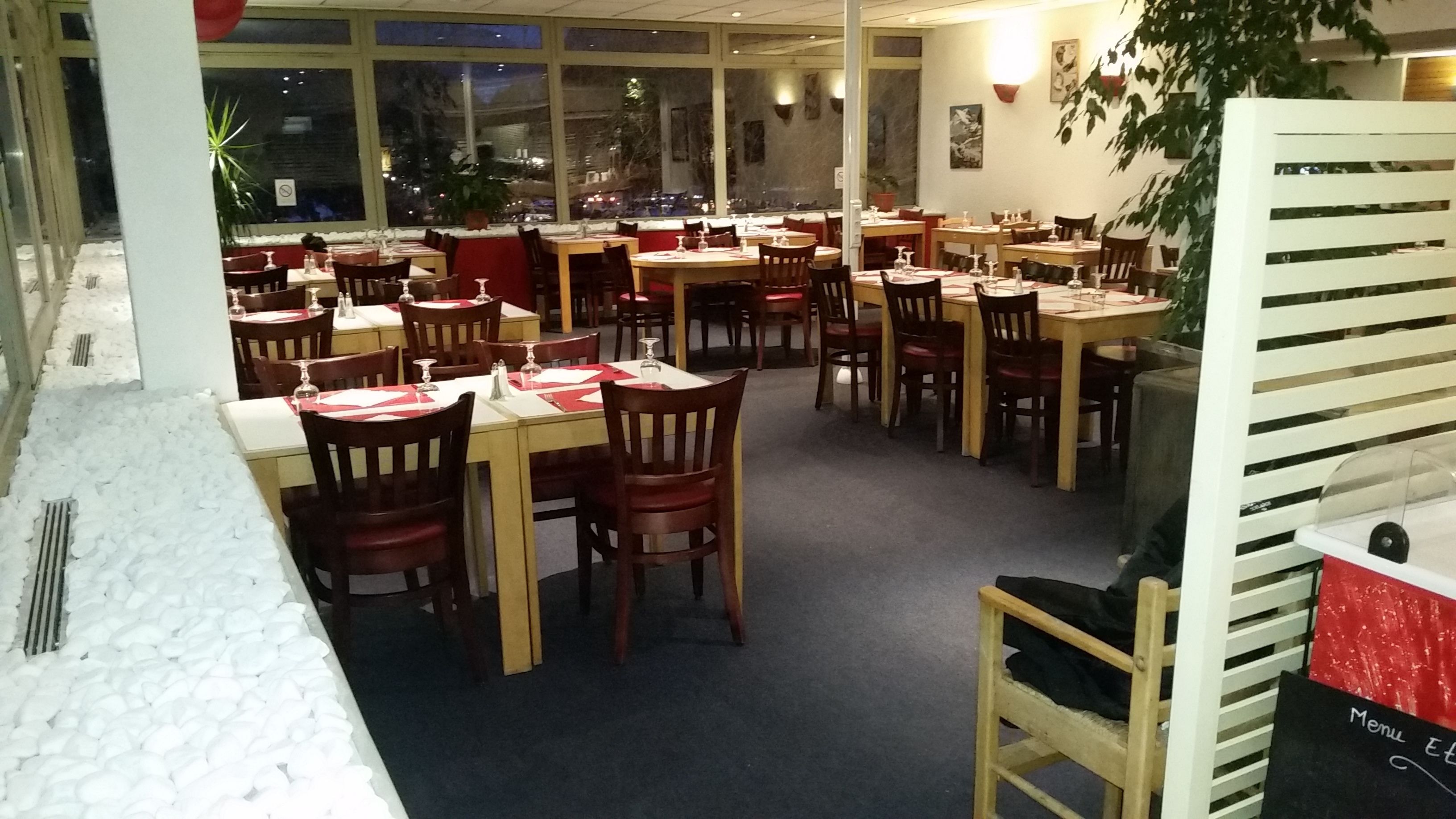 Een restaurant met meerdere tafels en stoelen, een menubord en een uitzicht op het exterieur door grote ramen.