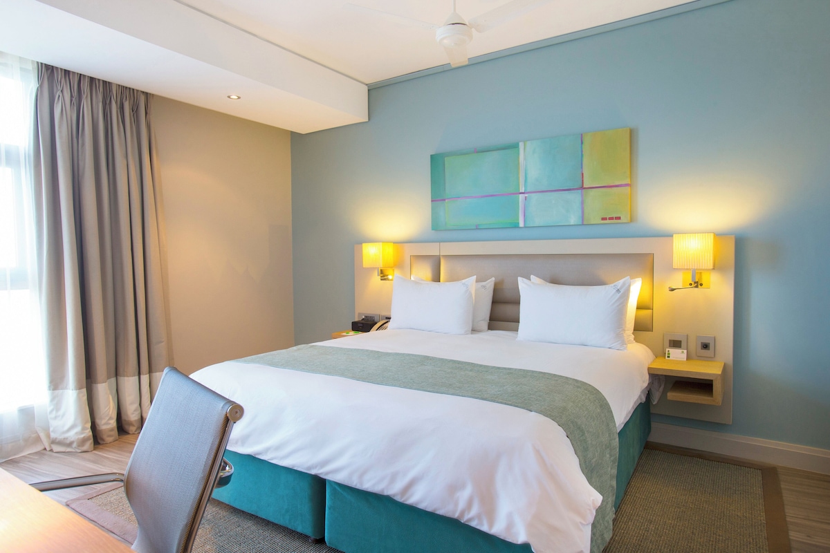 Quarto de hotel com uma cama grande, duas luminárias de cabeceira, cabeceira, mesa de cabeceira e uma janela com cortinas.