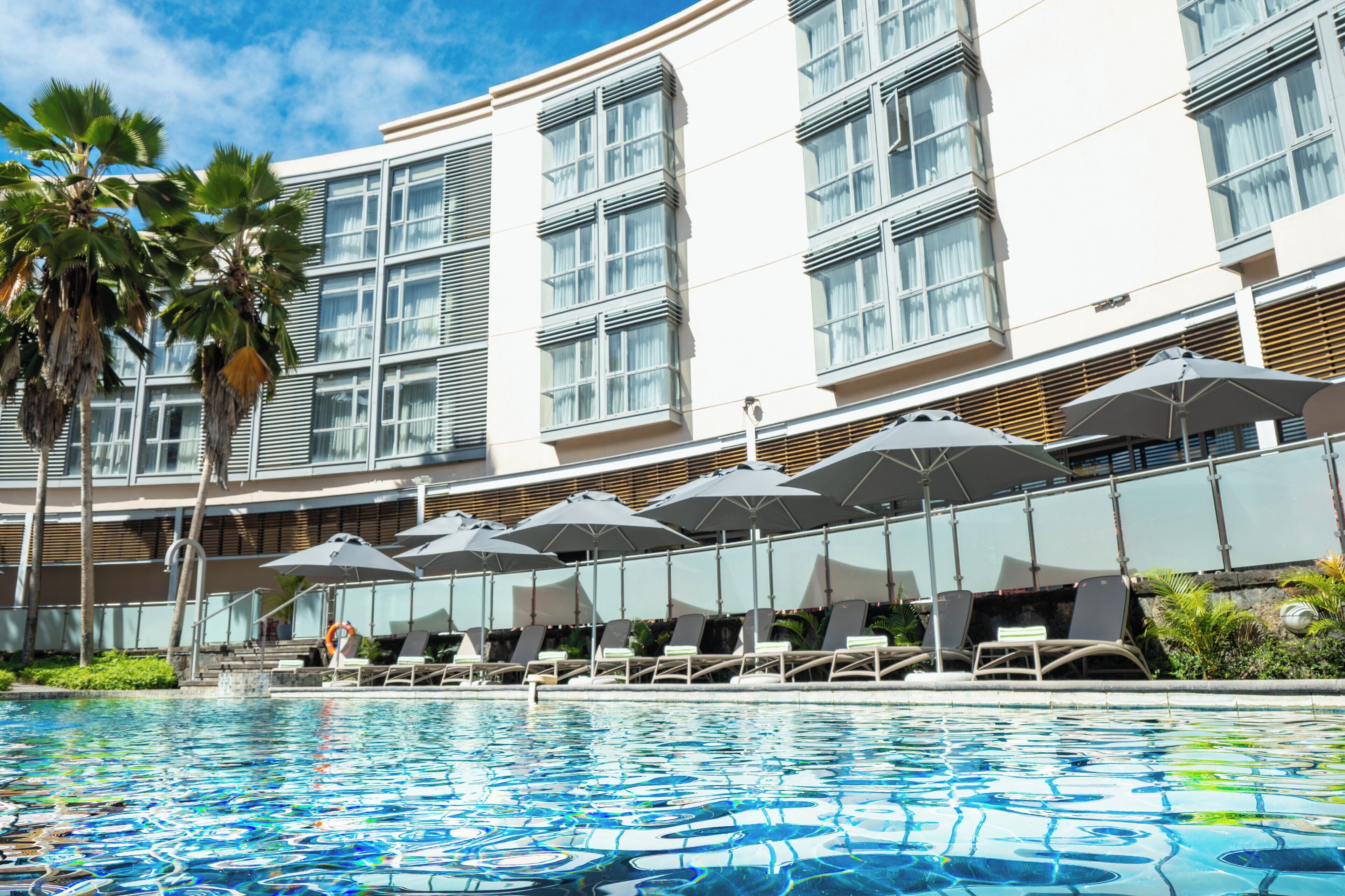 A hotel pool area with sun loungers and umbrellas, surrounded by a building with large windows and palm trees.