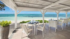 Un espace extérieur abrité, aménagé avec des tables et des chaises, offrant une vue sur une plage et l’océan.