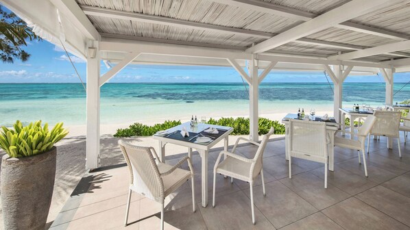 A covered outdoor dining area with tables and chairs, overlooking a beach and the ocean.