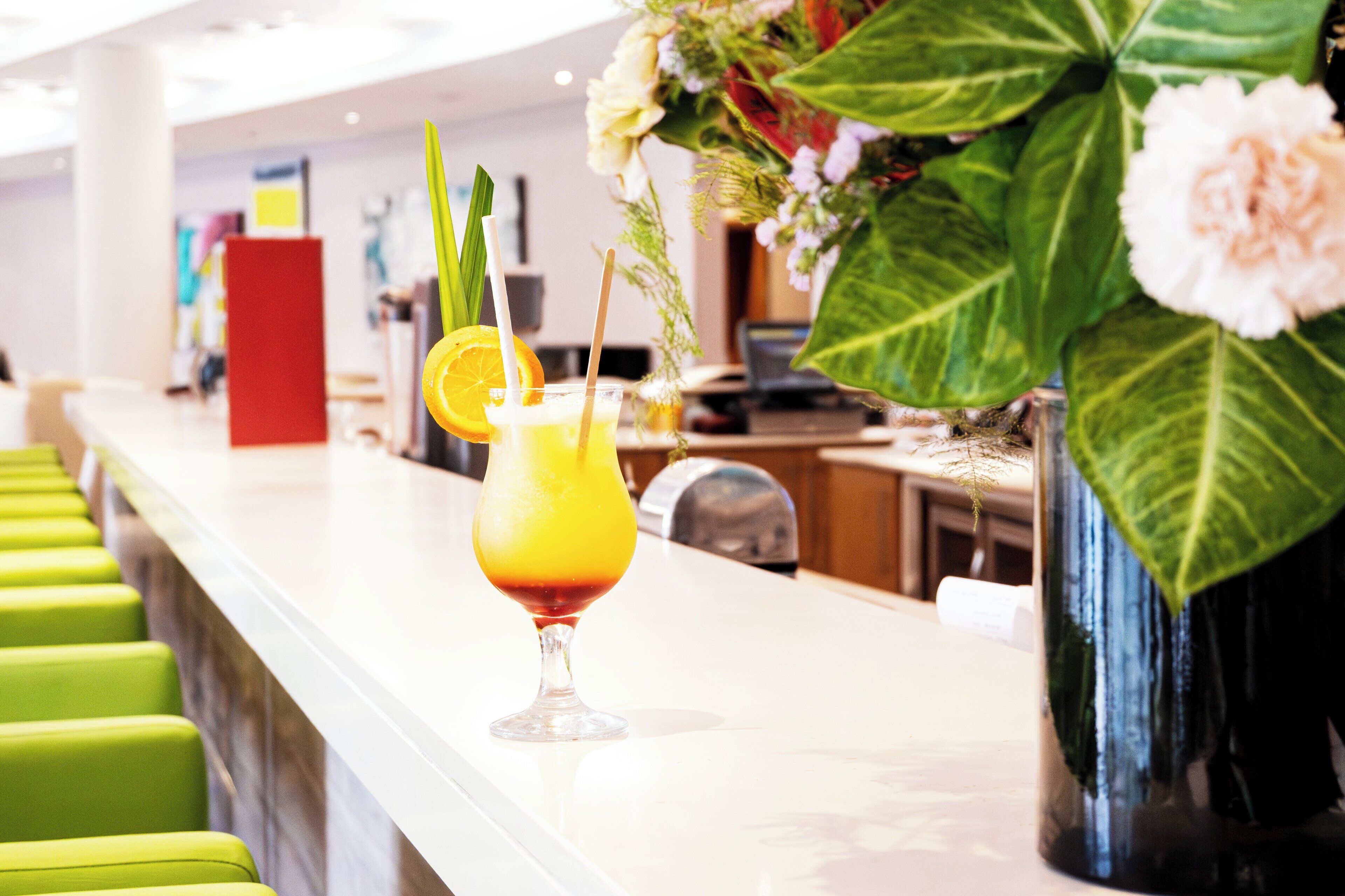 A bar area with a drink in a glass, green chairs, and a vase with flowers.
