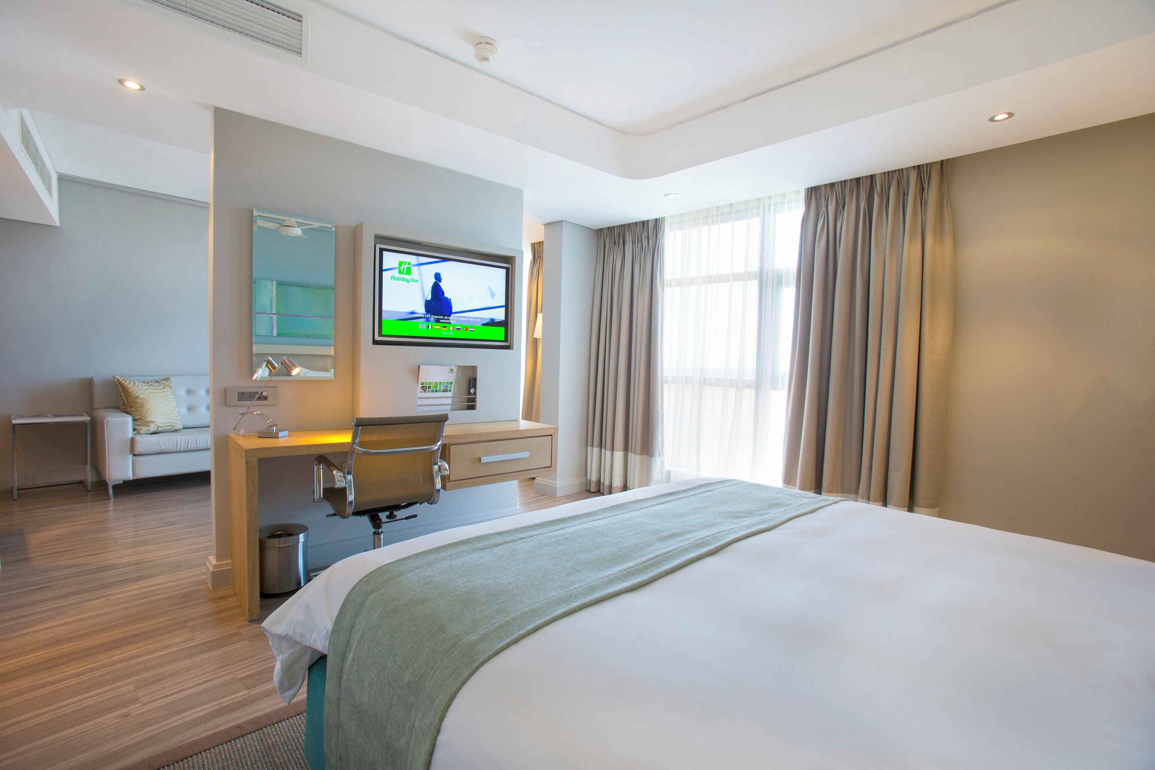 A modern hotel room with a large bed, a desk with a chair, a television, and a window with curtains.