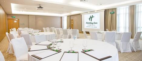 A conference room with round tables, white chairs, and a projector screen displaying 'Holiday Inn'.