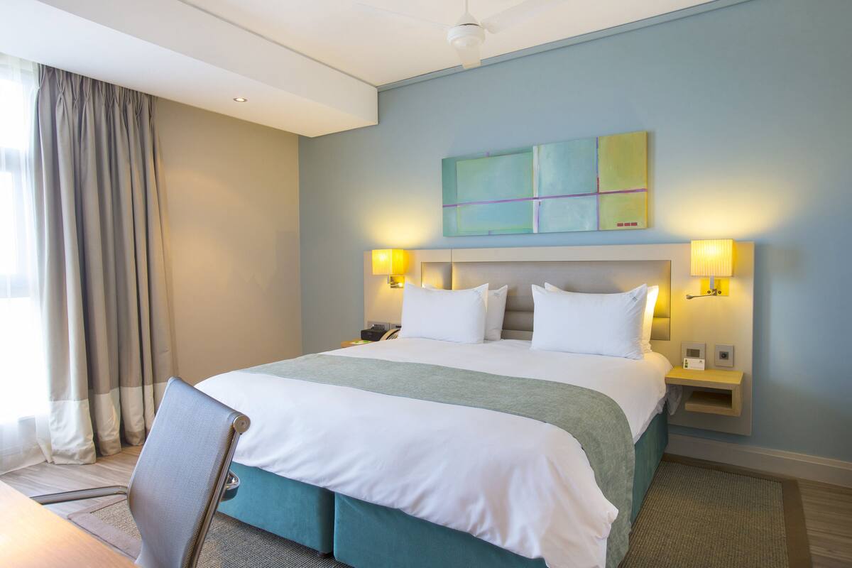 Une chambre d’hôtel avec un grand lit, deux lampes de chevet, une tête de lit, une table de chevet et une fenêtre avec des rideaux.